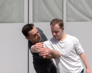 Zwei unterschiedliche Männer tanzen draussen vor einem Garagentor ein Duett. Zwei ihrer überlappenden Arme verbinden sich zu einem liegenden Oval.