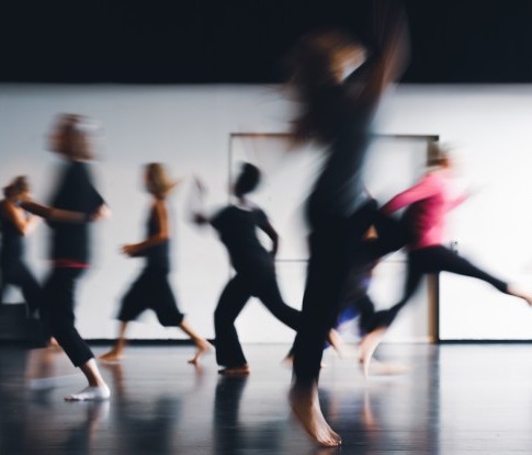 Eine Gruppe von Menschen durchquert mit sich kreuzenden Wegen ein Tanzstudio. Sie rennen, laufen und springen energievoll.